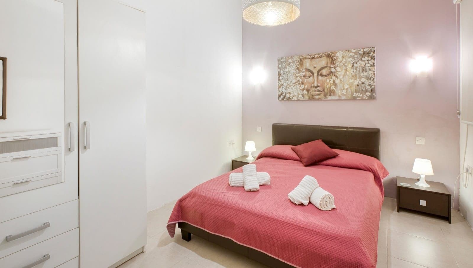 A hotel room with a bed, a nightstand, a lamp, a wall art piece, and a wardrobe.