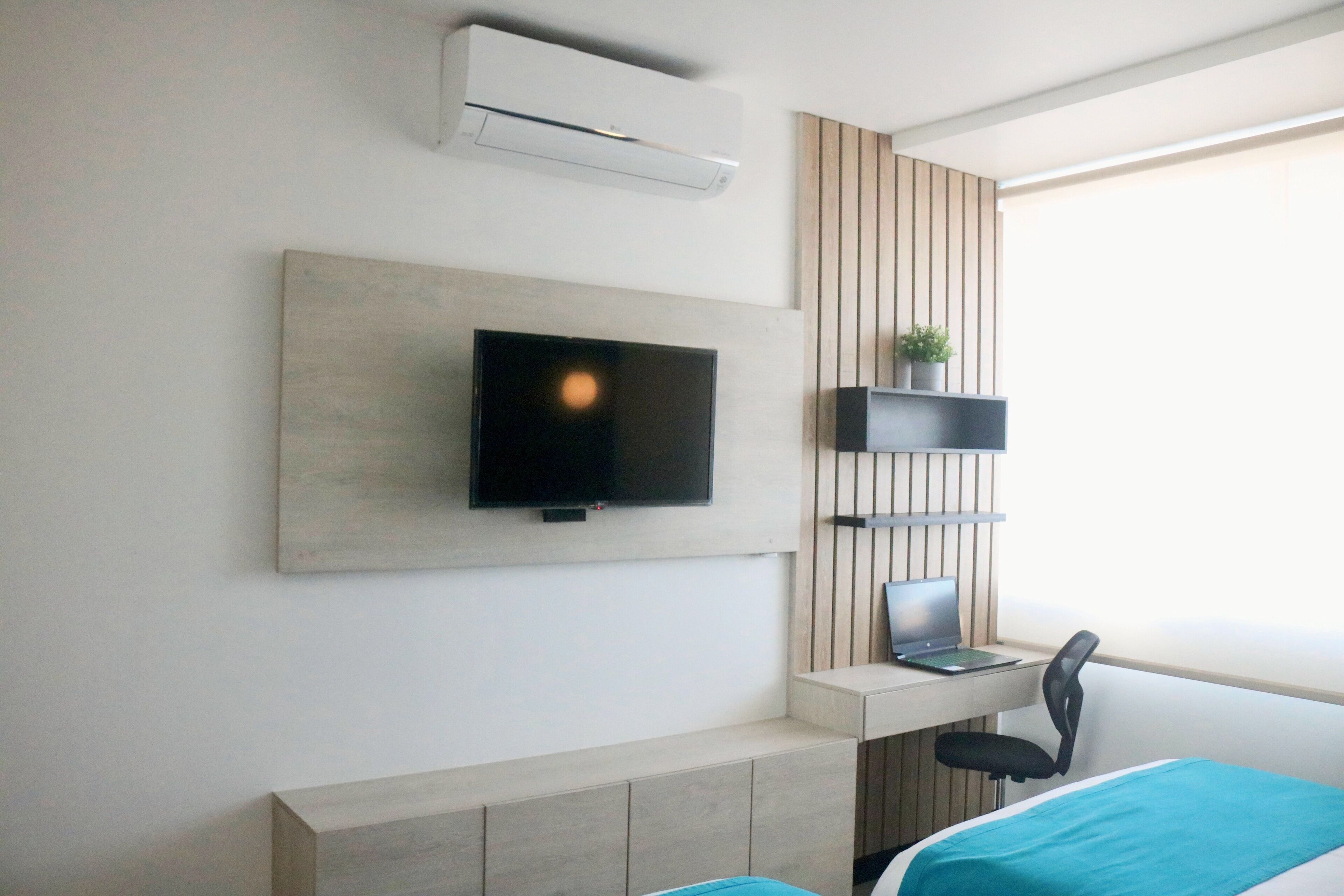 Una camera d'albergo moderna con TV a parete, una scrivania con un portatile, una sedia e un letto con copriletto blu.