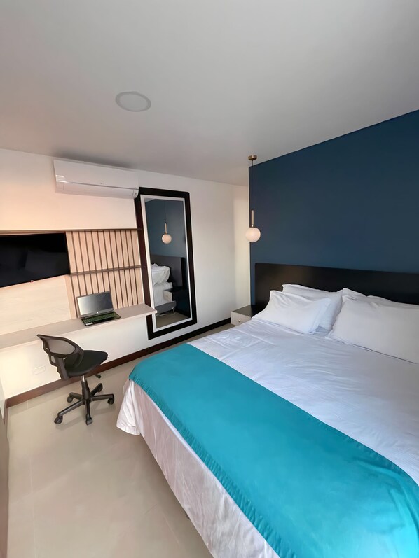 A modern hotel room with a large bed, a desk with a laptop, a mirror, and an air conditioning unit.