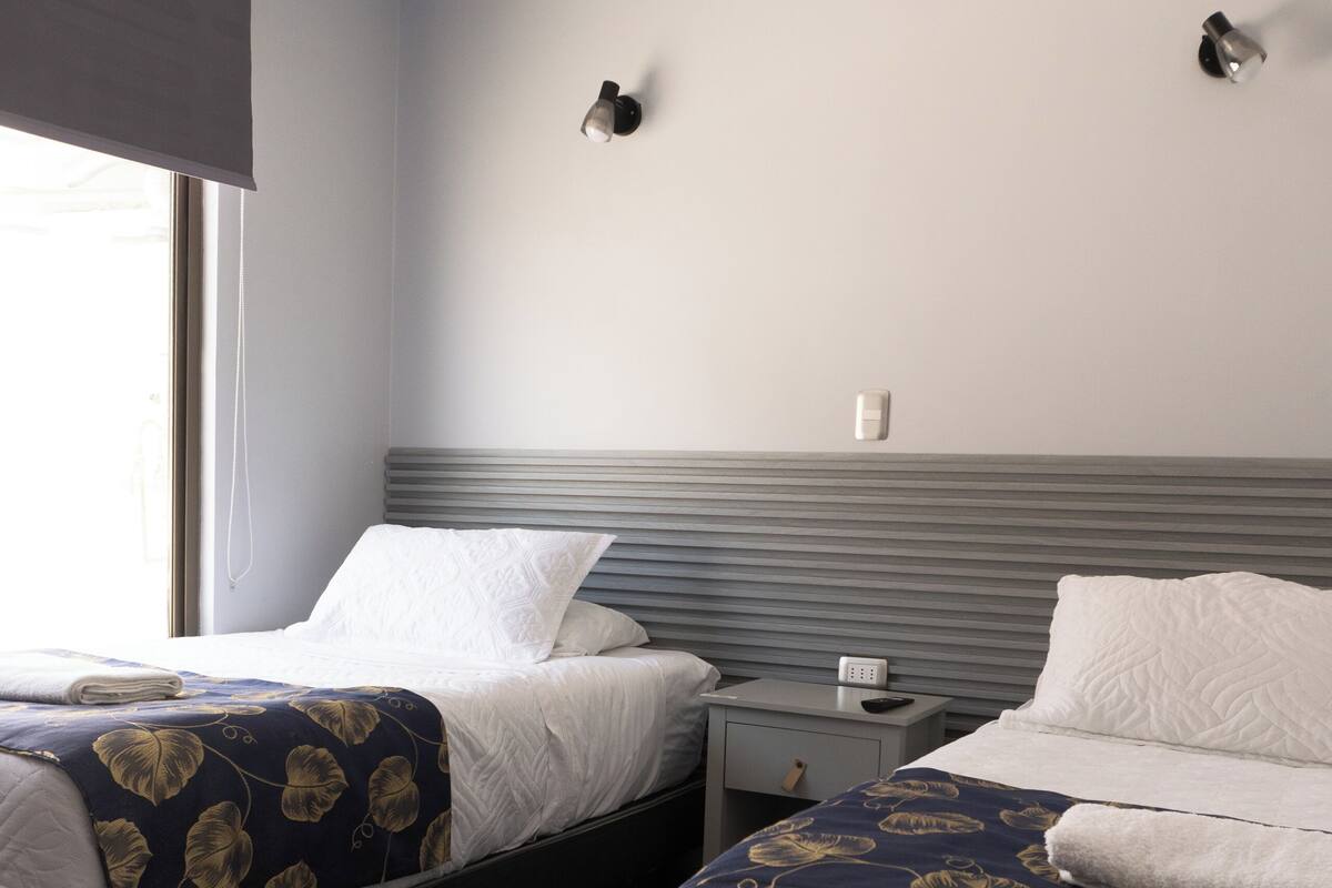 A hotel room with two beds, a bedside table with a lamp, and a window with a blind.