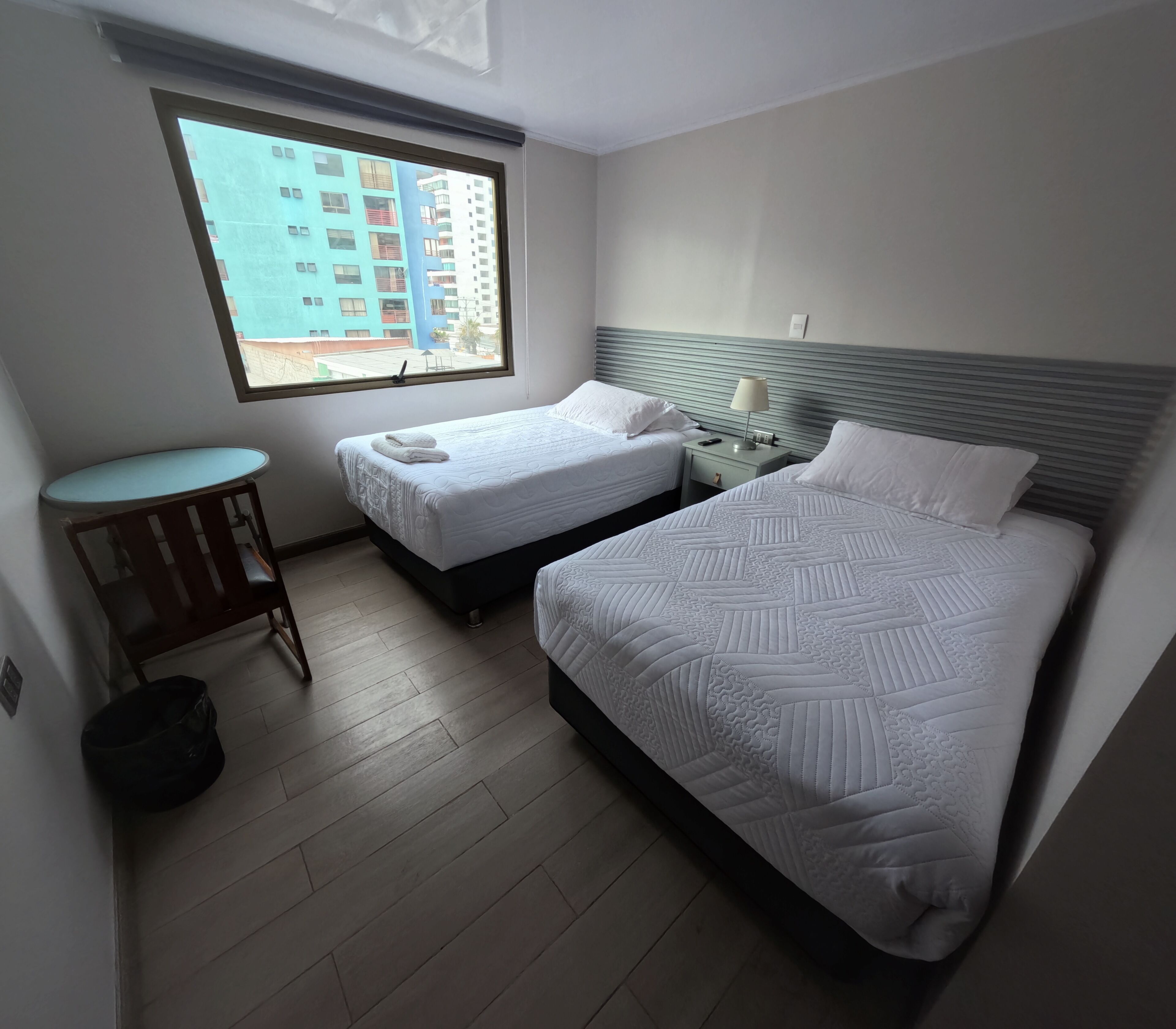 Habitación de hotel con dos camas, una mesita, una lámpara y una ventana con vista a edificios.