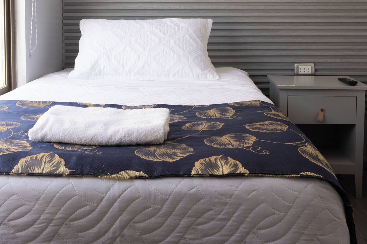 A neatly made bed with a patterned bedspread, a bedside table with a remote control, and a wall-mounted light fixture.