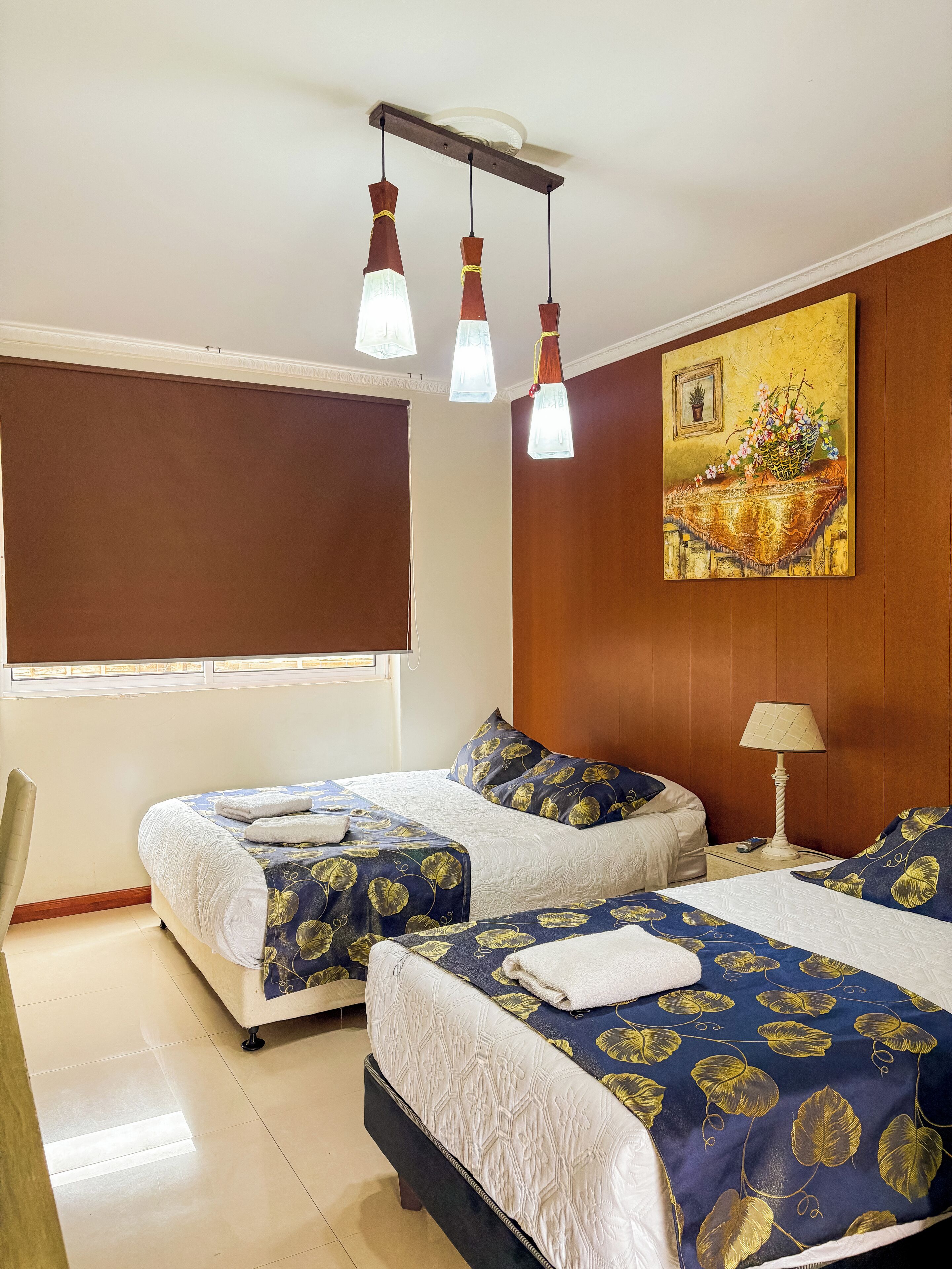Quarto de hotel com duas camas, um quadro na parede, uma janela com persianas e uma mesa de cabeceira com abajur.