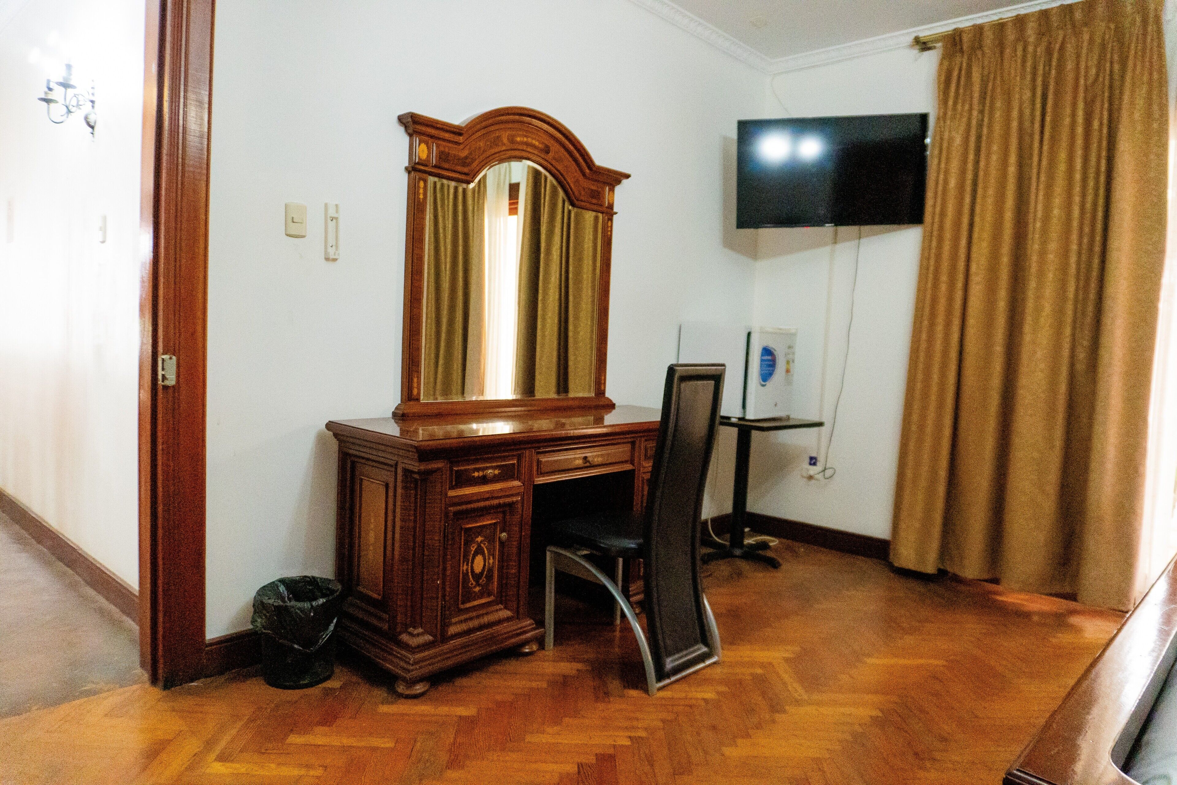 Una habitación con un escritorio de madera, una silla, un espejo grande, un televisor en la pared y una cortina.