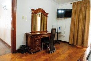 A room with a wooden desk, a chair, a large mirror, a television on the wall, and a curtain.