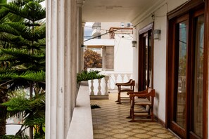 A balcony with a tiled floor, wooden chairs, a potted plant, and a view of a building in the background.