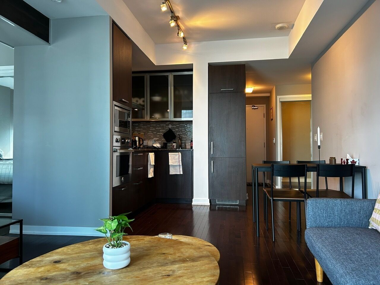 Een moderne woonkamer met een houten salontafel, een grijze bank en een eethoek met stoelen. Achtergrond toont een keuken met donkere kasten en een betegelde spatwand.