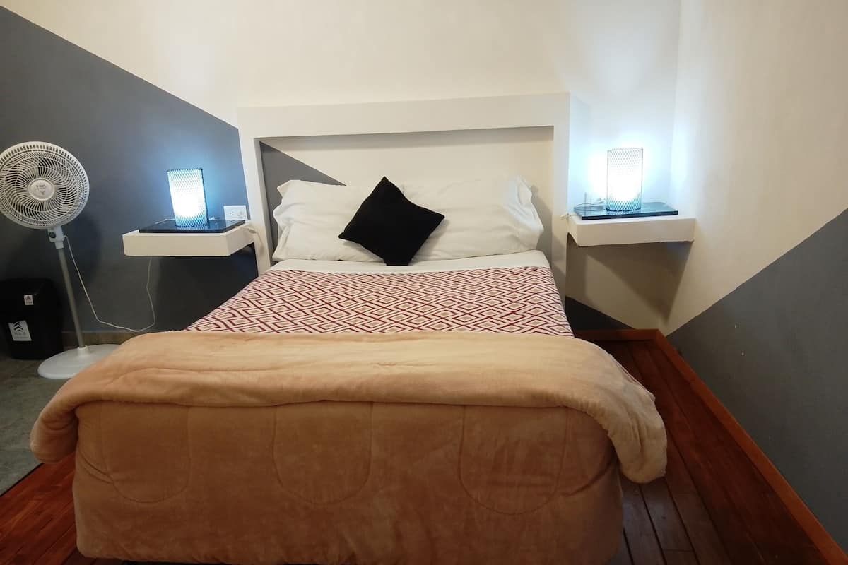 A neatly made bed with a patterned bedspread and a brown blanket, two bedside tables with lamps, a standing fan, and a small trash bin.