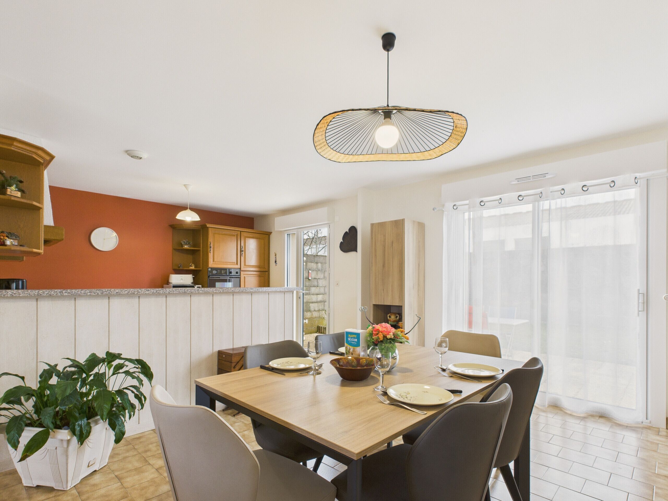 A dining area with a wooden table set for four, surrounded by chairs. There is a potted plant on the left and a vase with flowers on the table.