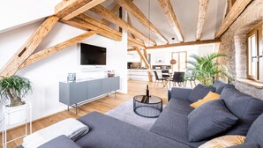 A modern loft apartment with exposed wooden beams, a grey sofa, a dining area, and a kitchen in the background. - Alte Schule Tiefenbach  (Östringen)
