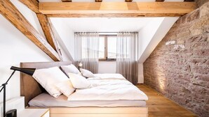 A bedroom with a sloped ceiling, exposed wooden beams, a stone wall, a bed with white bedding, and a bedside lamp. - Alte Schule Tiefenbach  (Östringen)