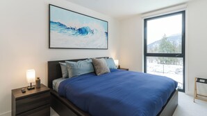 A bedroom with a large bed, a nightstand with a lamp, a window with a view of trees, and a painting on the wall. - The Luxury Basecamp (Revelstoke)