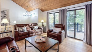 A spacious living room with a wooden ceiling, leather sofas, a coffee table, and large windows offering a view of the outdoors. - ROCK06 - Luxurious Sunriver Home, Hot Tub (Sunriver)