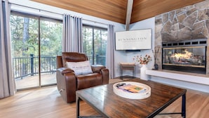 A modern living room with a leather recliner, a coffee table with books, a stone fireplace, and large windows offering a view of trees. - ROCK06 - Luxurious Sunriver Home, Hot Tub (Sunriver)