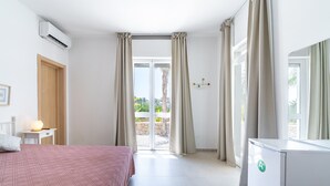 Habitación de hotel con cama, escritorio con lámpara, aire acondicionado, refrigerador y puerta corrediza de vidrio al exterior.