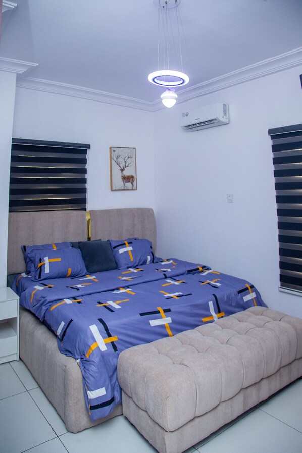 A bedroom with a bed, a bedside table, a wall-mounted air conditioner, and a ceiling light fixture. - Southern Village Serviced Apartments (Lekki)
