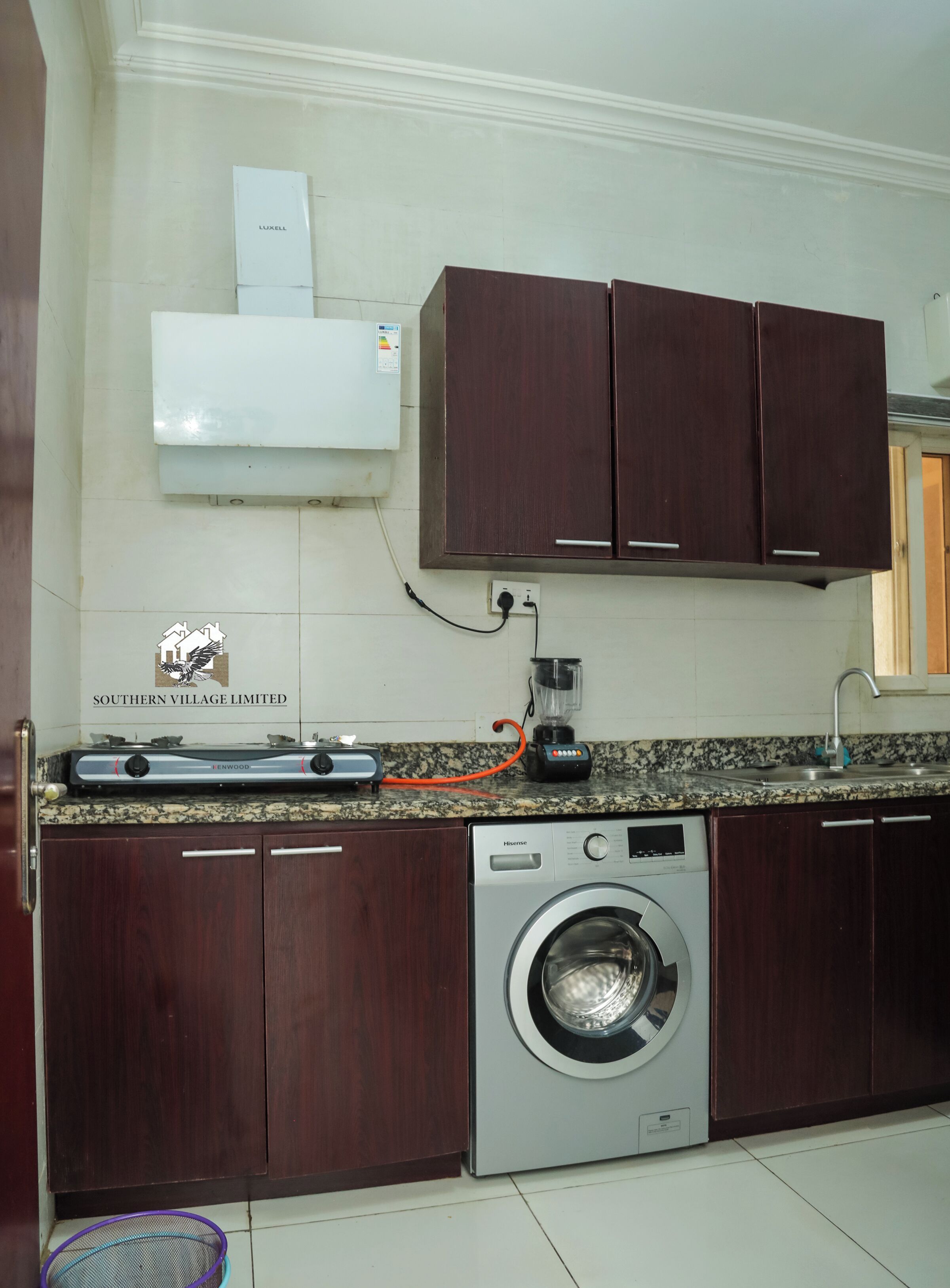 Une cuisine avec des meubles en bois foncé, un lave-linge encastré, un plan de travail avec un mixeur et une enseigne indiquant « Southern Village Limited ».
