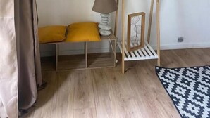 A room with a wooden floor, a bench with yellow cushions, a hanging lamp, a wooden rack with a basket, a framed picture, and a patterned rug. - Les Studios de la Côte Roz: Solo Traveler, Garden (Perros-Guirec)