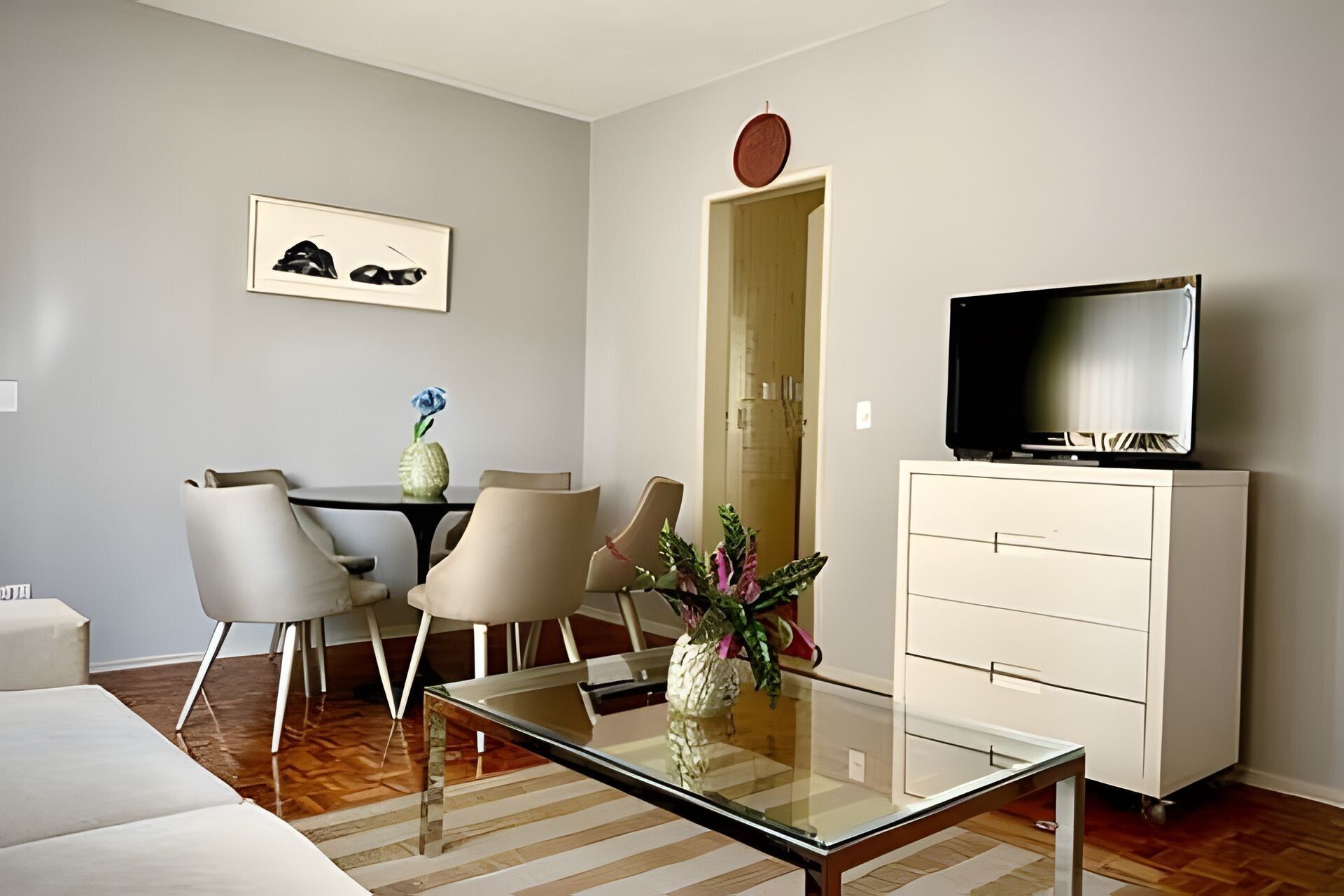 Una sala de estar moderna con una mesa de centro de vidrio, un sofá, un televisor sobre un mueble bajo, y un comedor con sillas y una mesa.