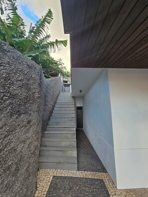 A narrow concrete staircase leading to a door, flanked by a stone wall and a modern building with a wooden roof. - Villa Barros (Câmara de Lobos)