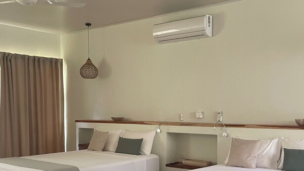 A hotel room with two single beds, a ceiling fan, an air conditioning unit, a bedside table with a lamp, and a large window with curtains.