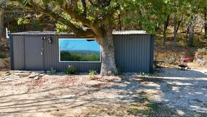 Une petite cabane à un seul étage, dotée d’une grande fenêtre et entourée d’arbres, avec un sol recouvert de gravier.