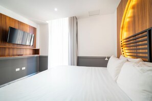 Quarto de hotel moderno com uma cama grande, televisão de tela plana presa a um painel de madeira, mesa de cabeceira e janela com cortinas.