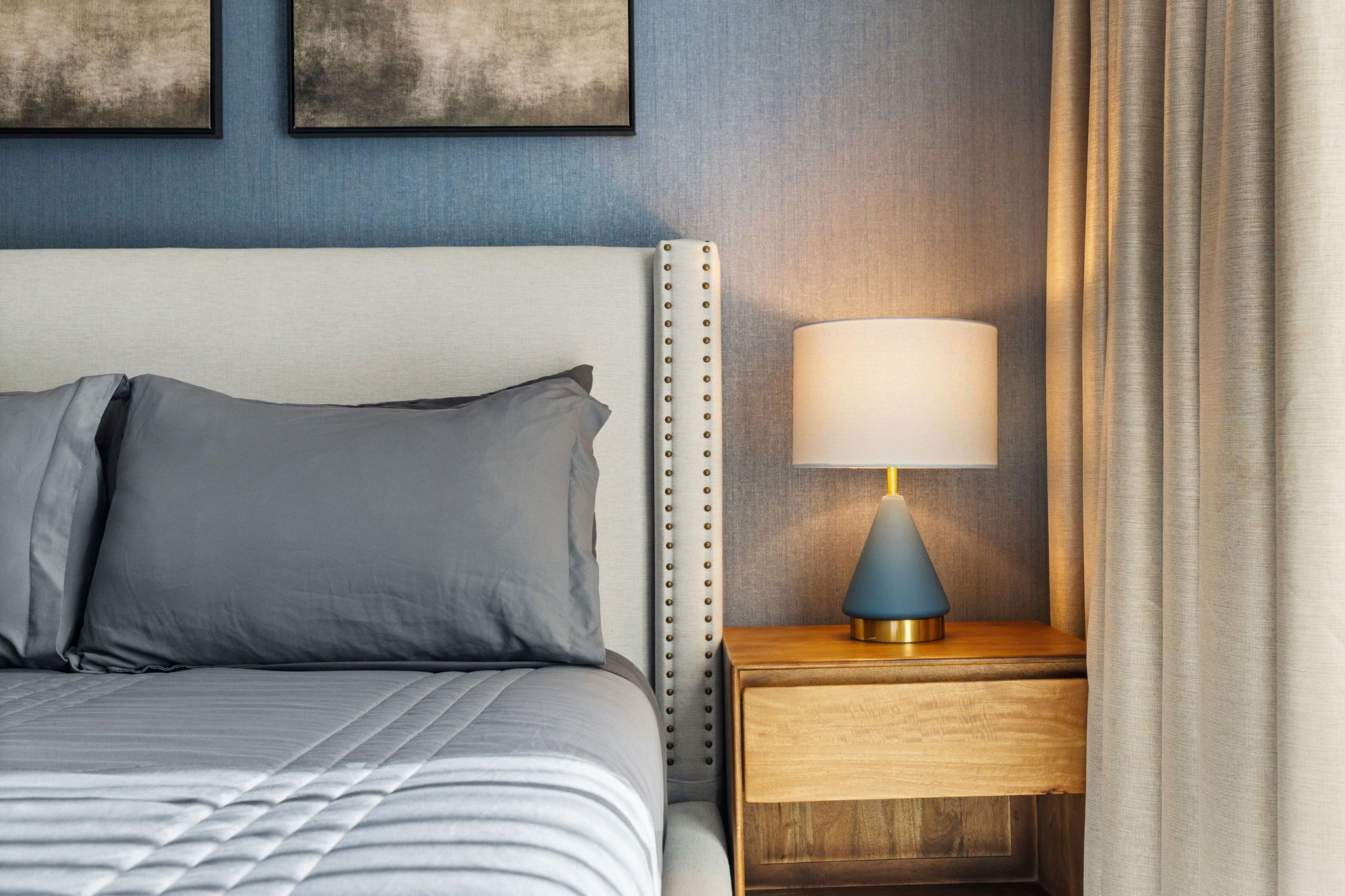 A neatly made bed with gray bedding, a wooden nightstand with a lamp, and a blue wall with artwork.