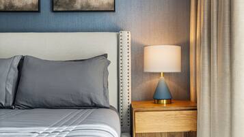 A neatly made bed with gray bedding, a wooden nightstand with a lamp, and a blue wall with artwork.