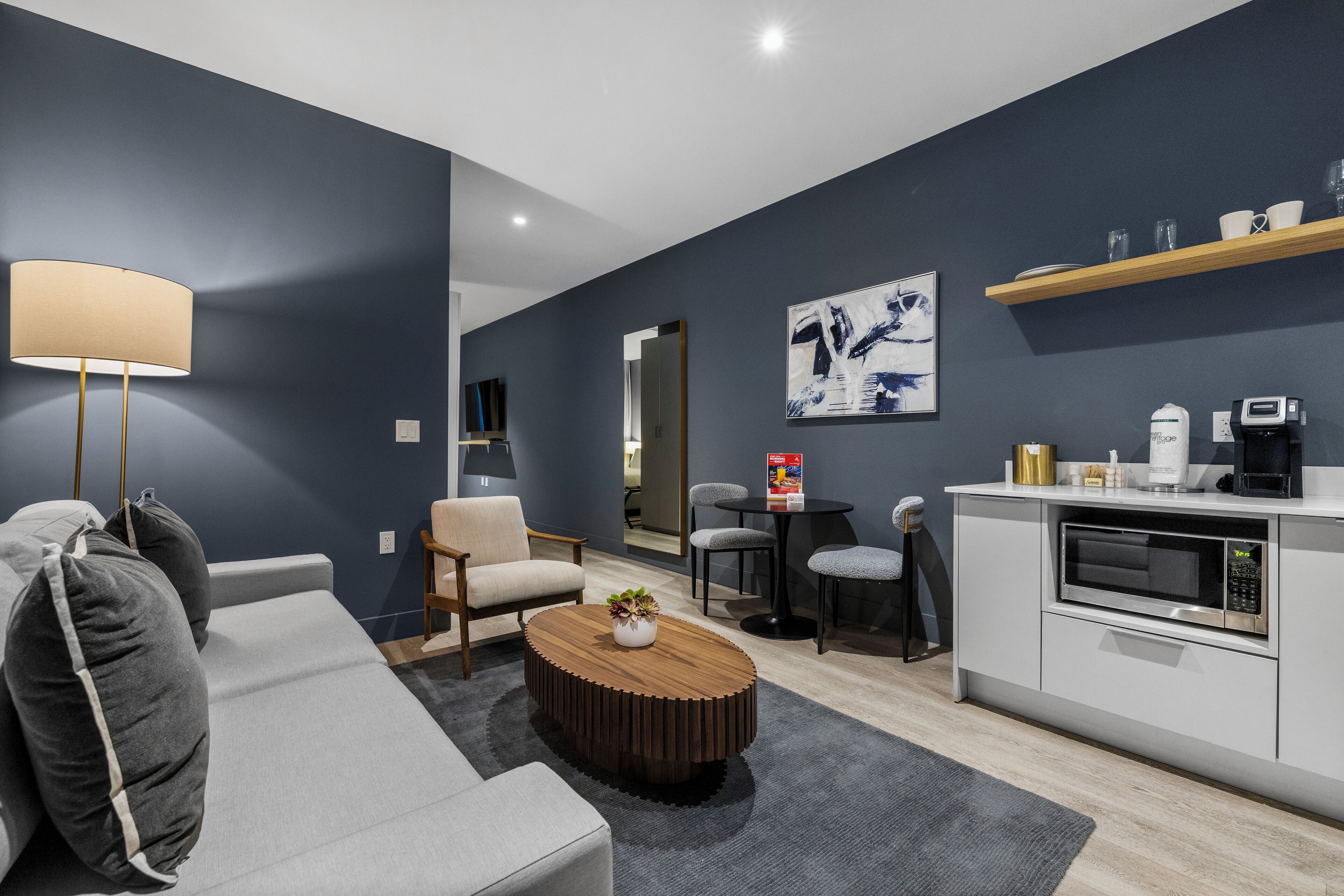 Ein modernes Wohnzimmer mit einer Couch, einem Couchtisch und Sesseln, einer Küchenzeile mit einer Mikrowelle sowie einem Esstisch mit Stühlen.