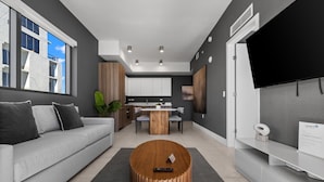 A modern living room with a grey sofa, a wooden coffee table, a dining area with a wooden table and chairs, and a flat-screen TV mounted on the wall. - Brickell Condos by Vibras Concierge (Miami)