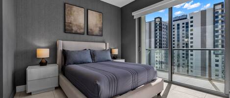 A modern bedroom with a large bed, two bedside tables, and a view of tall buildings through floor-to-ceiling windows.