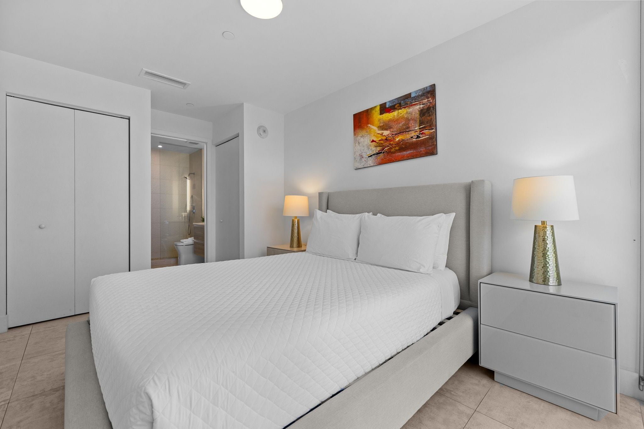 A modern bedroom with a large bed, two bedside tables with lamps, a door, and a painting on the wall.