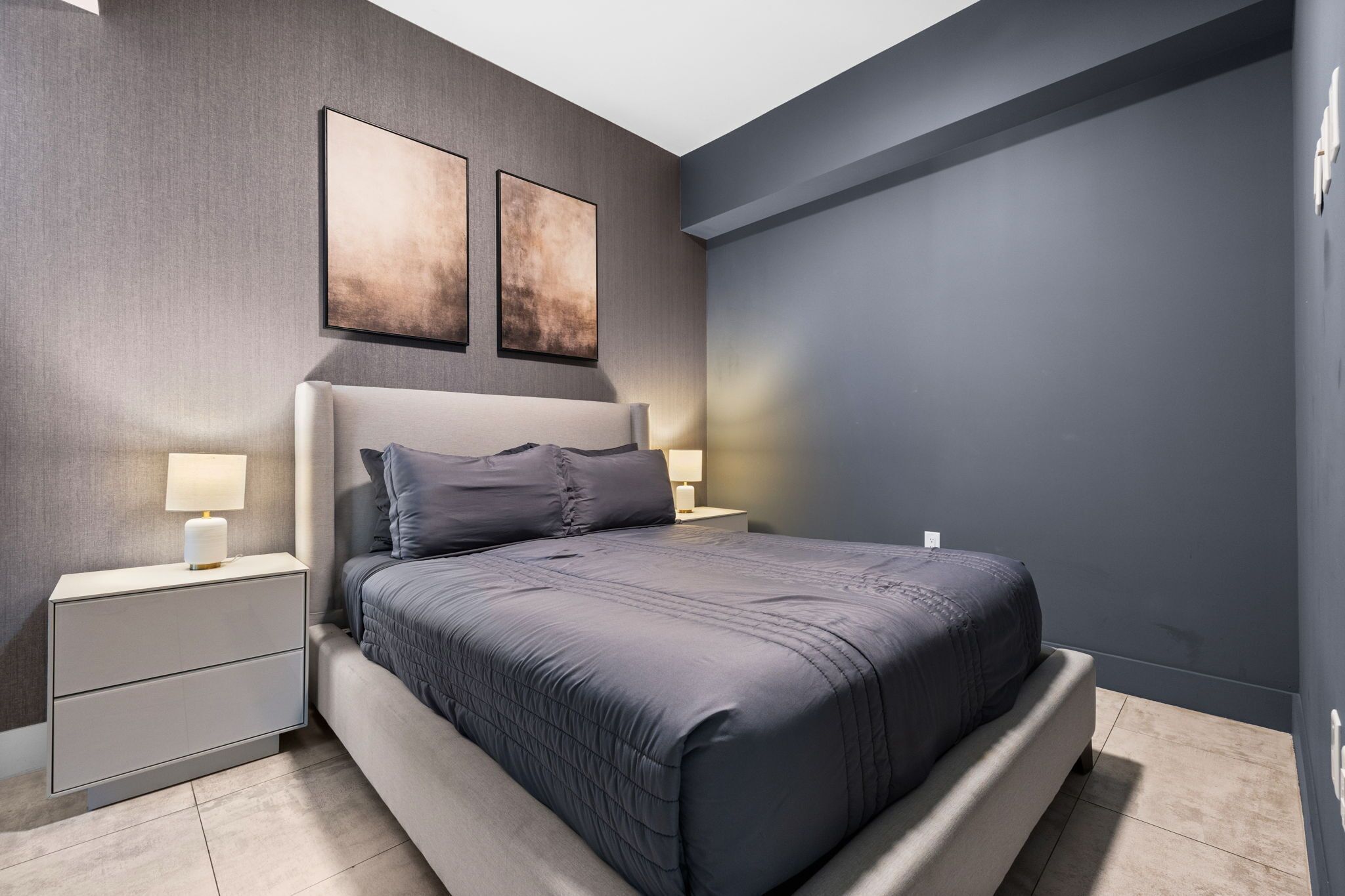 Une chambre à coucher moderne avec un grand lit, deux tables de chevet avec des lampes et deux tableaux abstraits accrochés au mur.