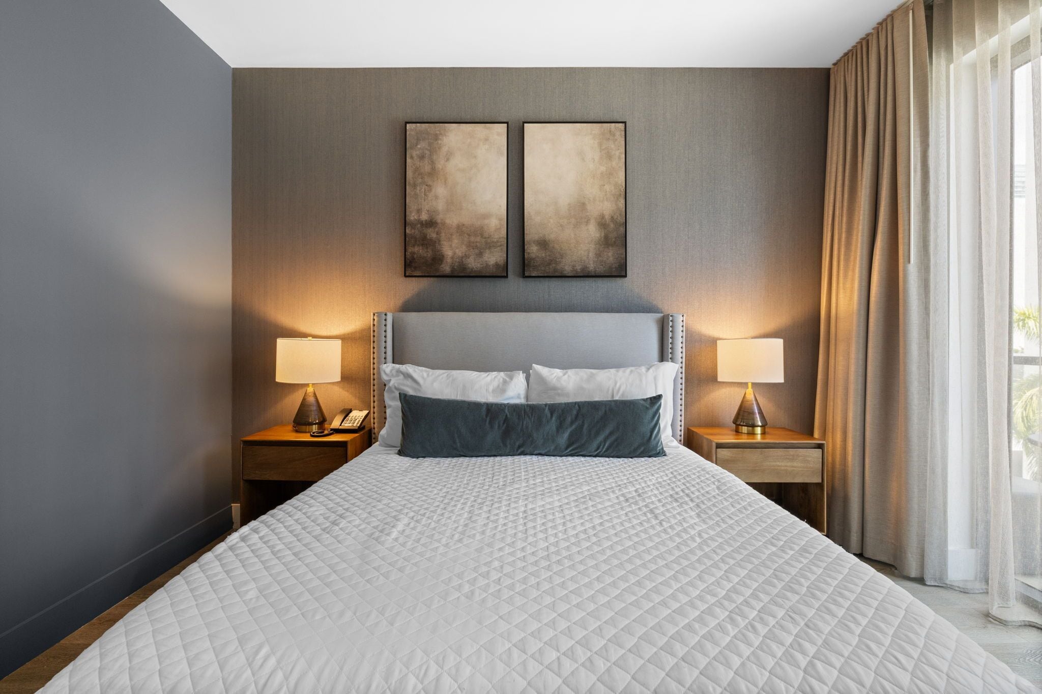 A hotel room with a large bed, two bedside tables with lamps, and two abstract wall art pieces above the bed.