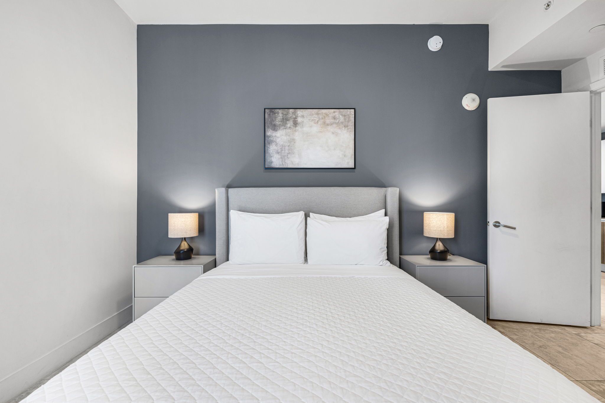 Un dormitorio moderno con una cama grande, dos mesitas de noche con lámparas, una pared de acento gris y una puerta.