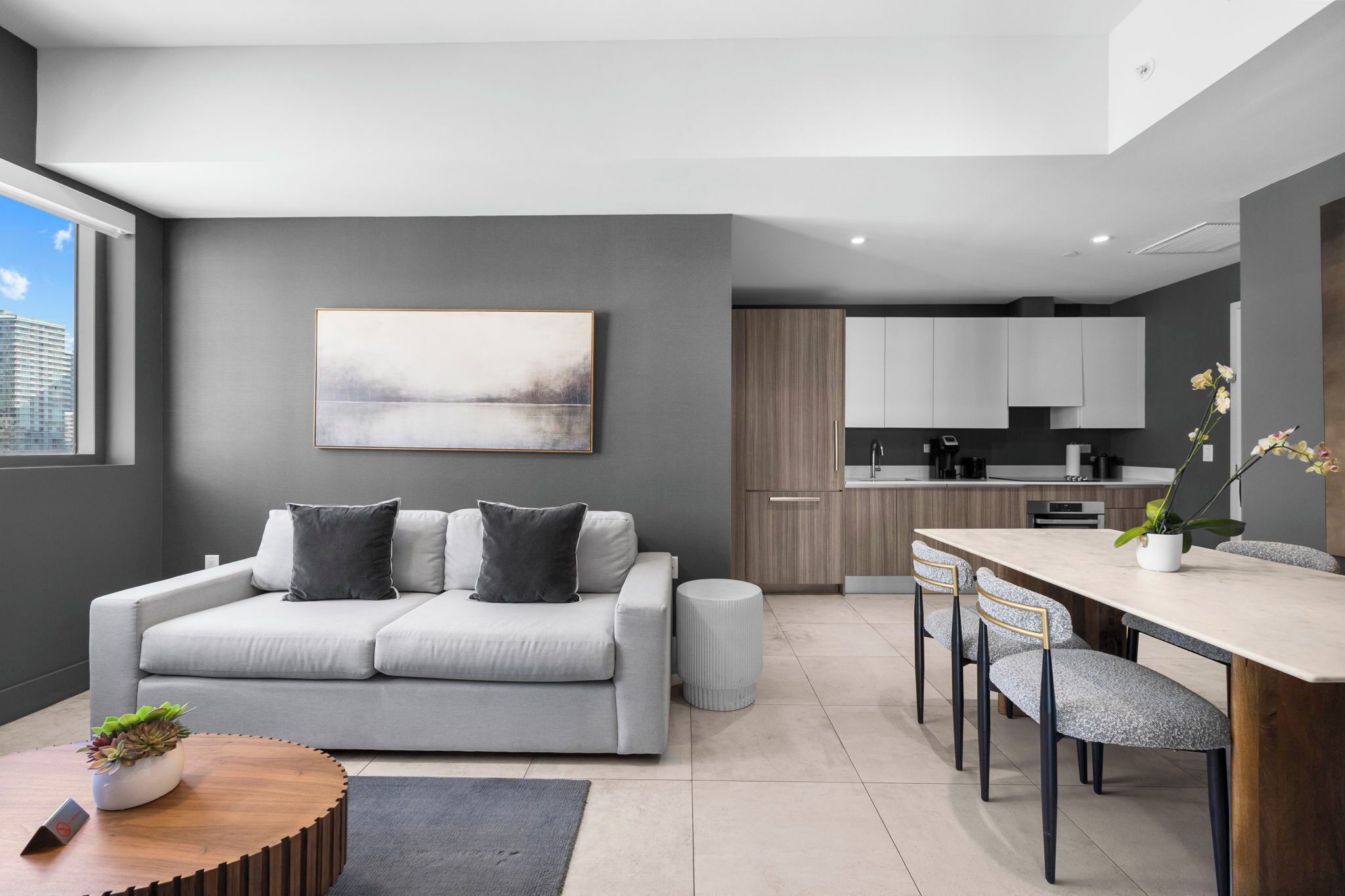 Una sala moderna con un sofá gris, una mesa de centro con una planta, y un comedor con una mesa y sillas de madera.