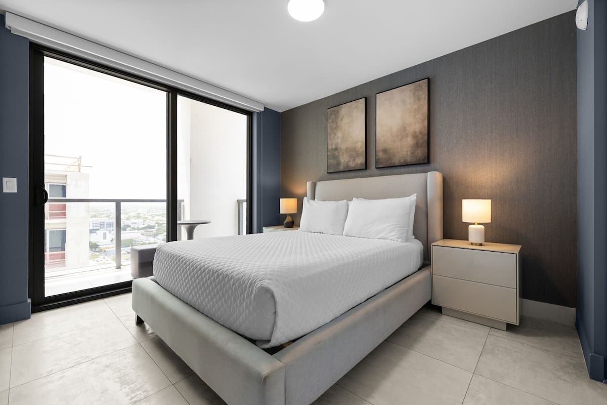 A modern hotel bedroom with a large bed, two bedside tables with lamps, a view through a sliding door, and artwork on the wall.
