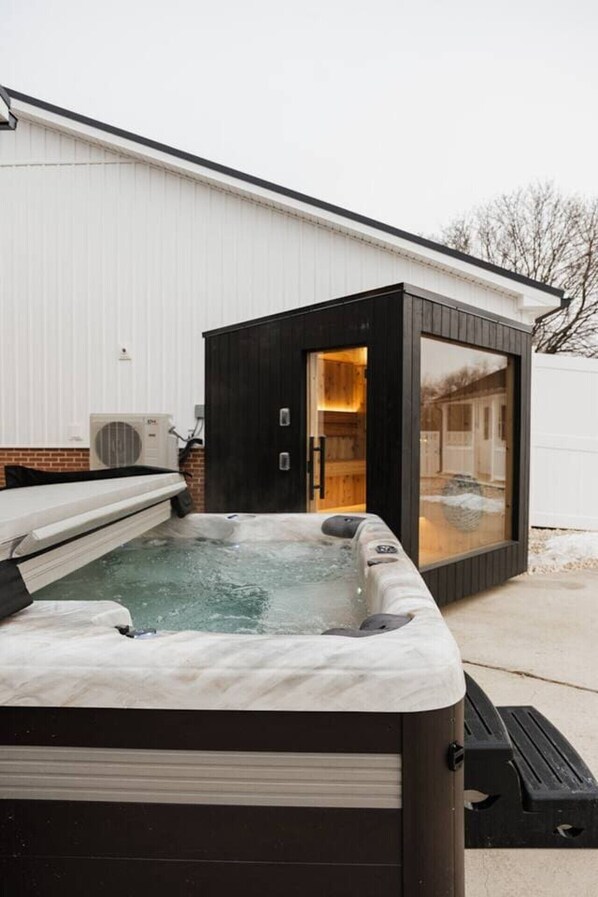 Ein Whirlpool mit Holzterrasse, eine kleine Holzhütte mit Glasüren und ein weißes Gebäude im Hintergrund.