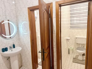 A bathroom with a wooden door, a white sink with blue cups, a mirror with lights, and a toilet with a patterned cover. - The best 5bedroom house with AC in a vibrant yet low key part of Accra. (Accra)