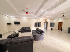 A spacious living area with a large sectional sofa, a smaller sofa, and a coffee table. There are ceiling fans, a flat-screen TV mounted on the wall, and a door leading to another room. - The best 5bedroom house with AC in a vibrant yet low key part of Accra. (Accra)