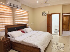 A bedroom with a bed, bedside tables, an air conditioner, a ceiling fan, a bathroom door, and a tiled floor. - The best 5bedroom house with AC in a vibrant yet low key part of Accra. (Accra)