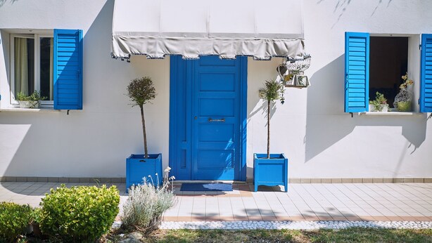 Een gebouw met een blauwe deur, witte muren en blauwe luiken. Aan weerszijden van de deur staan potplanten.