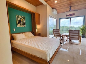 A bedroom with a bed, a nightstand, a window, and a view of greenery outside. - Serenity of Nature - Tiny Home in Manuel Antonio (Quepos)