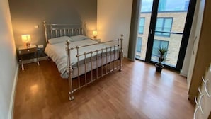 Un dormitorio con cama de estructura metálica, dos mesitas de noche con lámparas, un amplio ventanal con vistas a edificios y una planta en maceta.