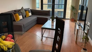 Una sala moderna con un sofá gris, una mesa de centro de vidrio y una mesa de comedor con sillas. Hay una planta en maceta y un ventanal con vista a un edificio.