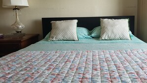 Una cama con colcha floral, dos almohadas estampadas con cuadros y una mesita de noche con lámpara.