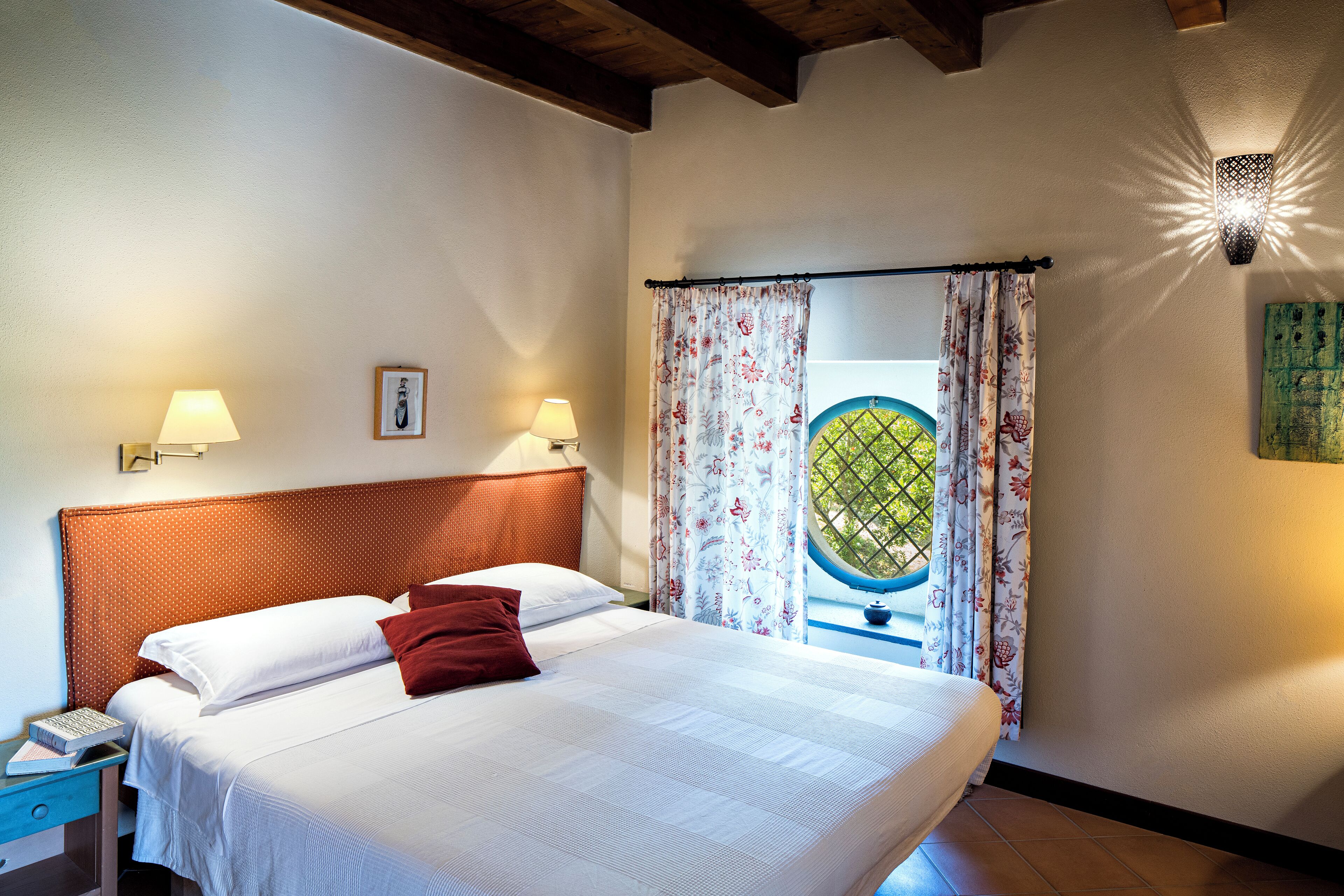 A neatly made bed with white linens and a single red pillow, flanked by two bedside lamps. A framed picture hangs on the wall above the bed.
