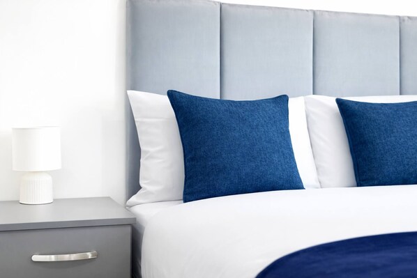 A neatly made bed with a grey headboard, white and blue pillows, and a white bedside lamp on a grey nightstand with a drawer. - Work-friendly 1-bed Leicester (Leicester)