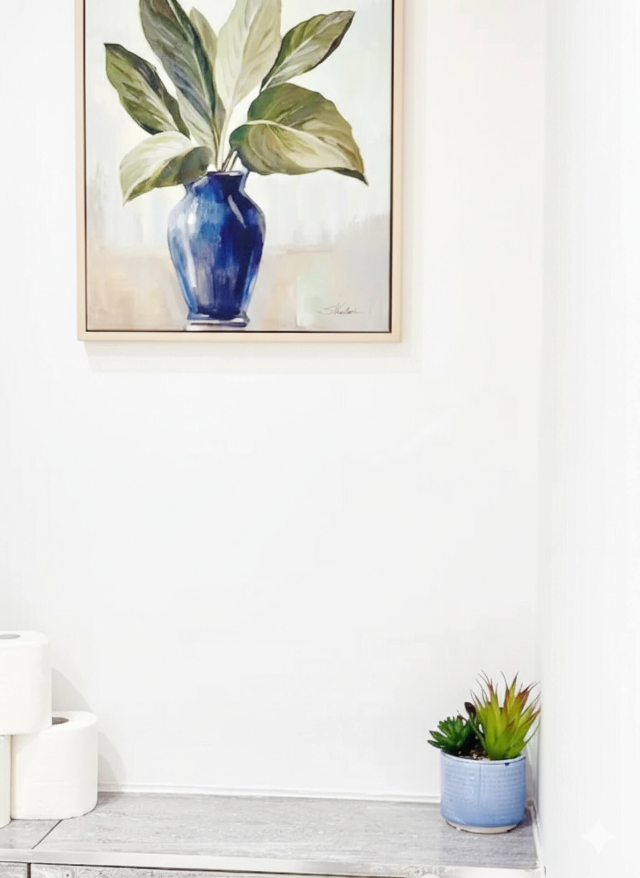 A framed painting of a blue vase with green leaves on a white wall, a small potted plant on a shelf, and a roll of toilet paper on the floor.